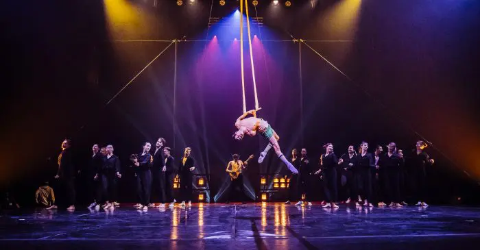 Un artiste aérien en costume rose est suspendu à des sangles jaunes, au centre de la scène lors des 2022 Spectacles annuels de l'ENC, entouré d'un groupe de personnes en tenues sombres sous un éclairage théâtral dramatique.