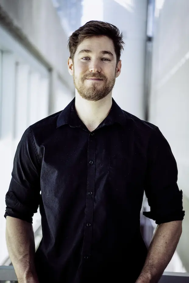 Ashley Youren, un homme barbu aux cheveux bruns courts, vêtu d'une chemise à boutons sombre aux manches retroussées, se tient à l'intérieur devant un arrière-plan flou et moderne, souriant légèrement à l'appareil photo.
