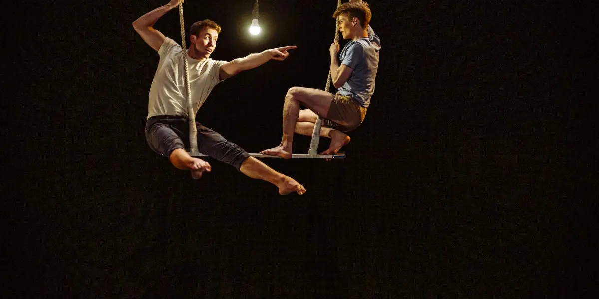 Deux acrobates, Maxime Charron et Corentin Lemaître Auger, se balancent dans les airs sous une seule ampoule suspendue - l'un pointant vers l'extérieur, l'autre assis - sur un fond sombre.
