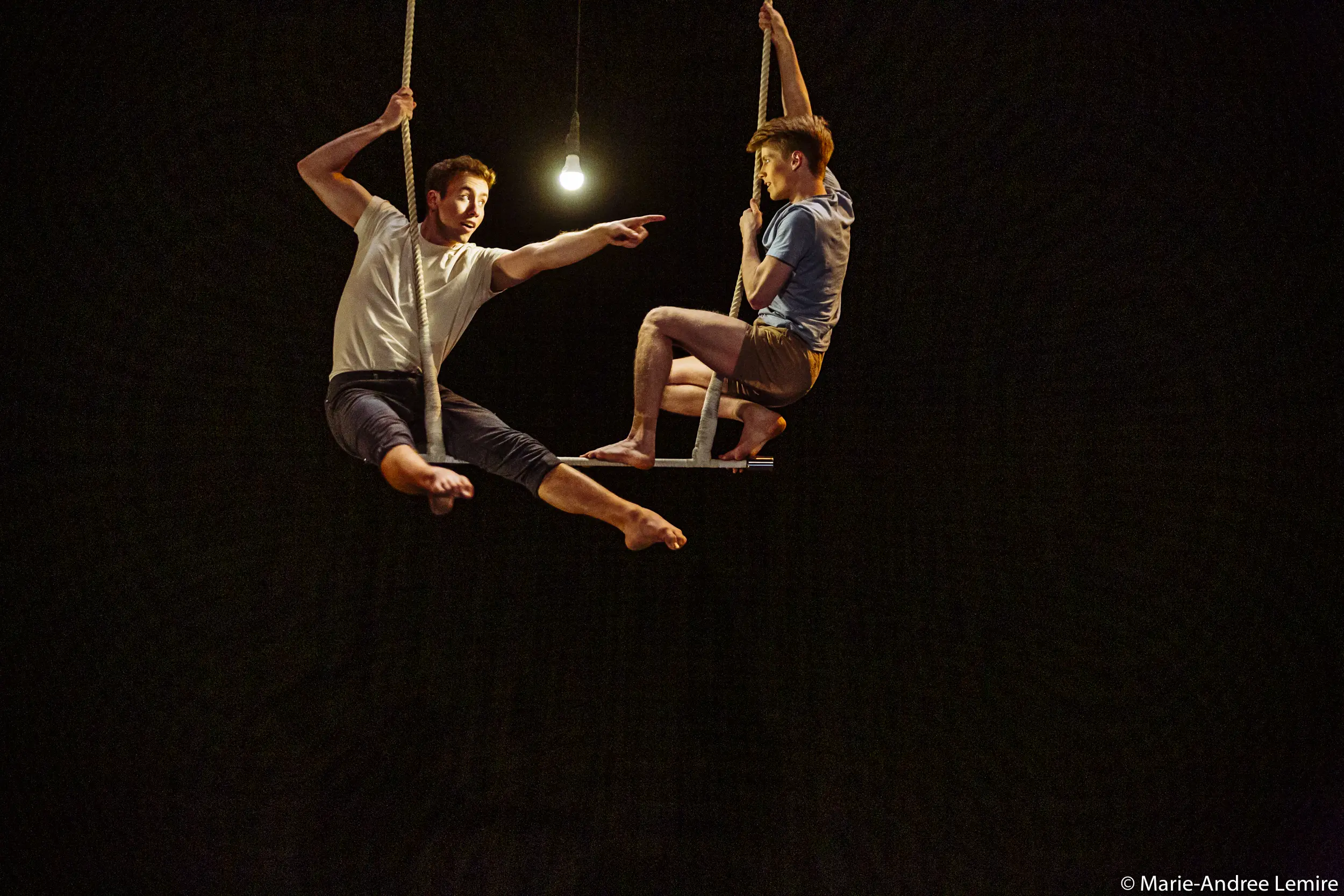 Deux acrobates, Maxime Charron et Corentin Lemaître Auger, se balancent dans les airs sous une seule ampoule suspendue - l'un pointant vers l'extérieur, l'autre assis - sur un fond sombre.