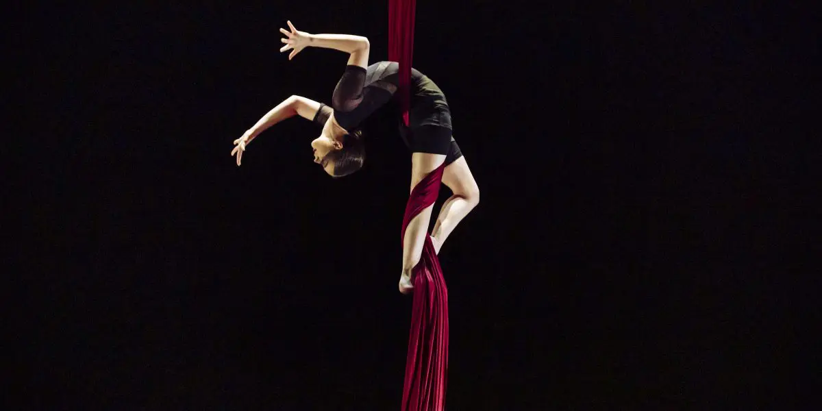Une voltigeuse, Jasmine Gagné-Morin, vêtue d'une tenue noire, exécute une élégante pose en flexion arrière dans les airs, suspendue à des soies rouges sur un fond sombre.
