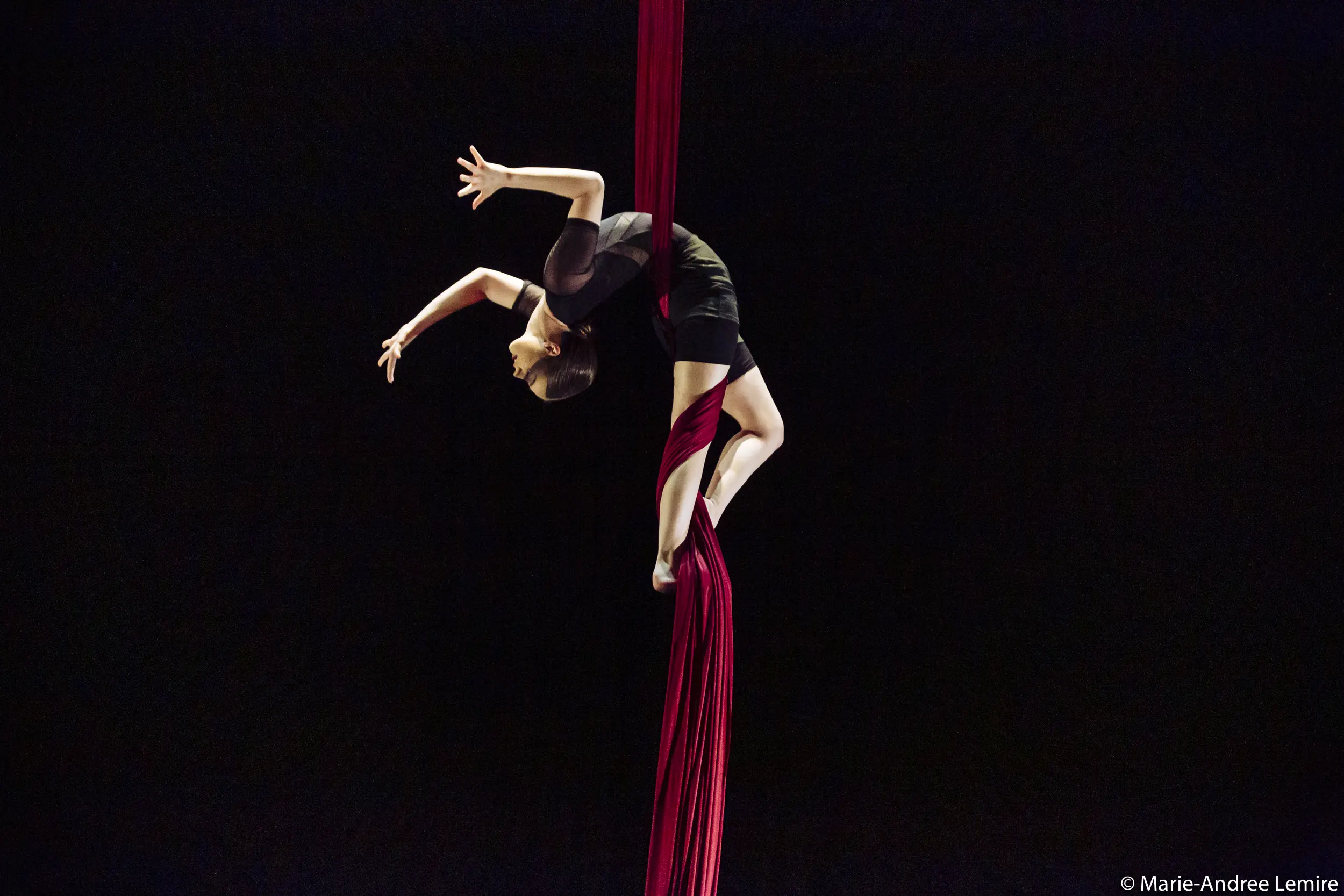 Une voltigeuse, Jasmine Gagné-Morin, vêtue d'une tenue noire, exécute une élégante pose en flexion arrière dans les airs, suspendue à des soies rouges sur un fond sombre.