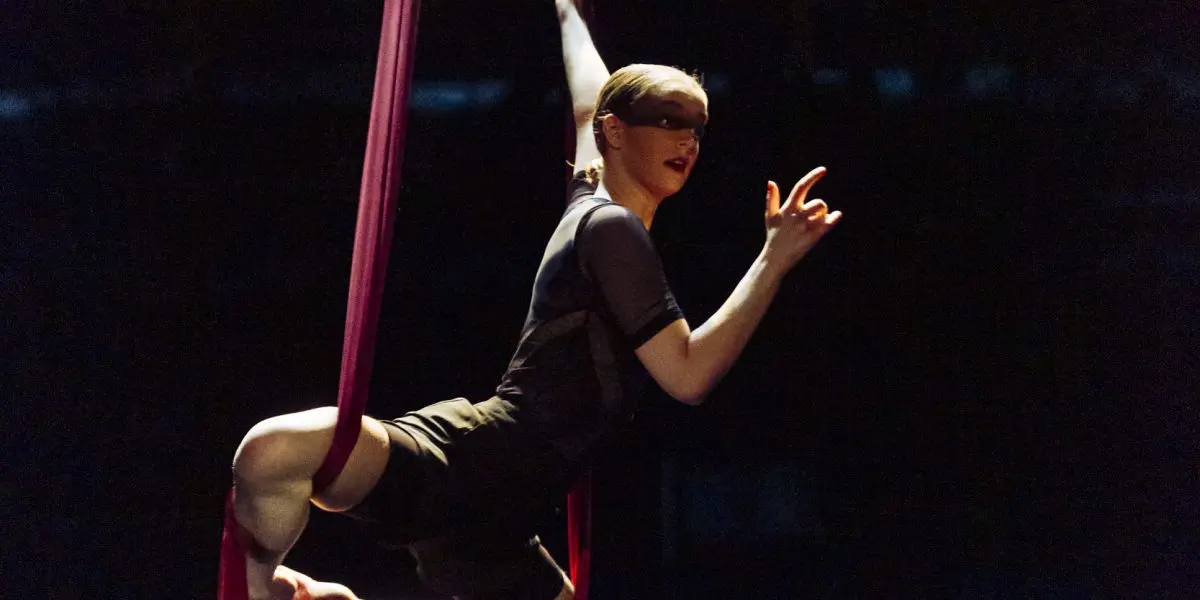 Une voltigeuse, Jasmine Gagné-Morin, se produit sur de la soie rouge, suspendue dans les airs avec une jambe enveloppée de tissu et un bras tendu, sur un fond de scène sombre.