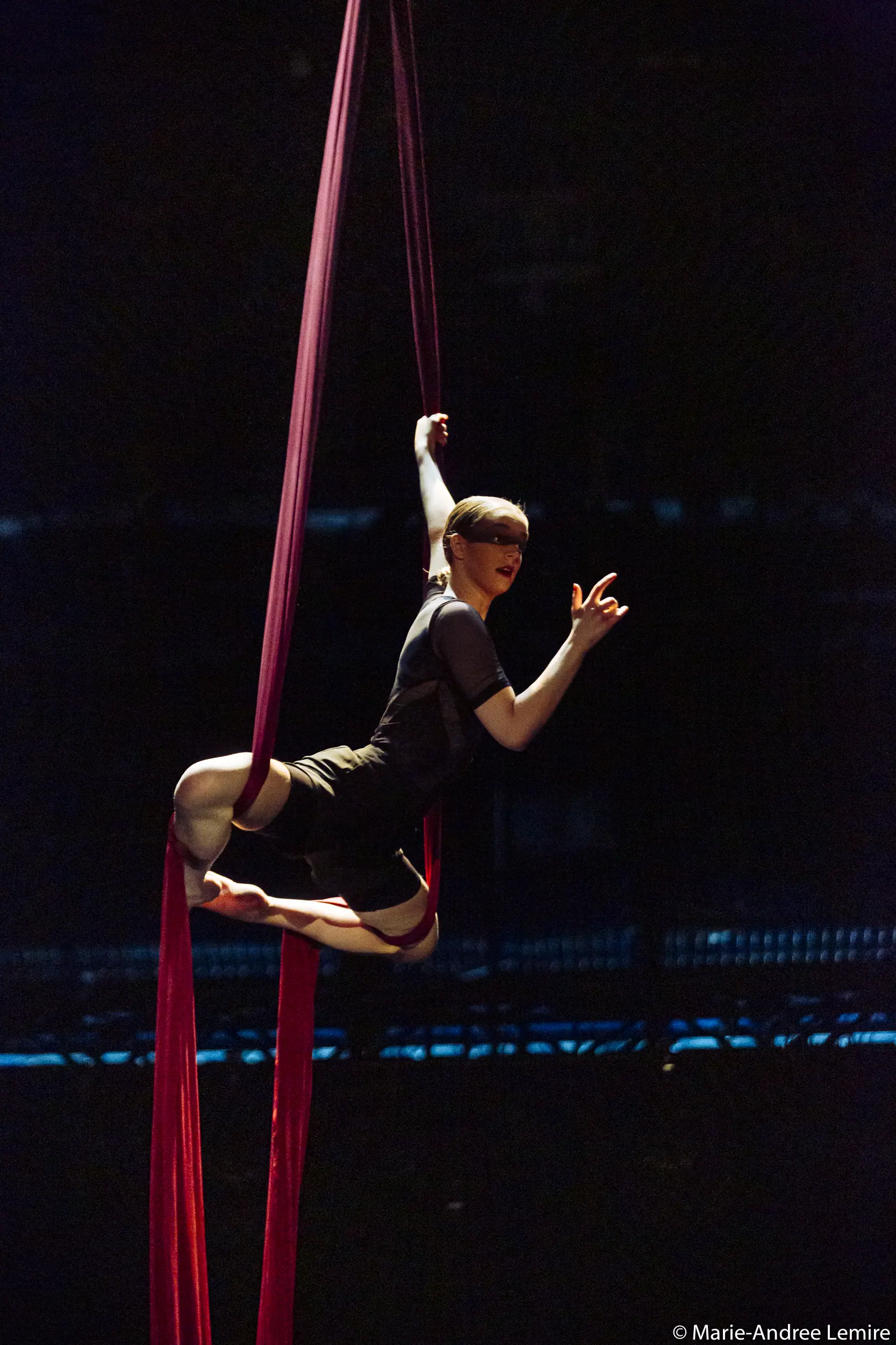 Une voltigeuse, Jasmine Gagné-Morin, se produit sur de la soie rouge, suspendue dans les airs avec une jambe enveloppée de tissu et un bras tendu, sur un fond de scène sombre.
