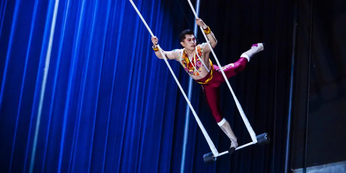 L'artiste de cirque Gabriel Olmos Cruz se tient gracieusement en équilibre sur un trapèze, un bras tenant une corde, une jambe tendue, sur fond de rideaux d'un bleu profond.