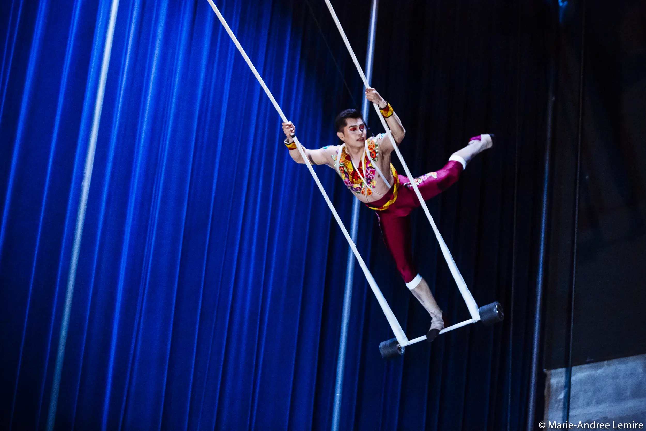 L'artiste de cirque Gabriel Olmos Cruz se tient gracieusement en équilibre sur un trapèze, un bras tenant une corde, une jambe tendue, sur fond de rideaux d'un bleu profond.