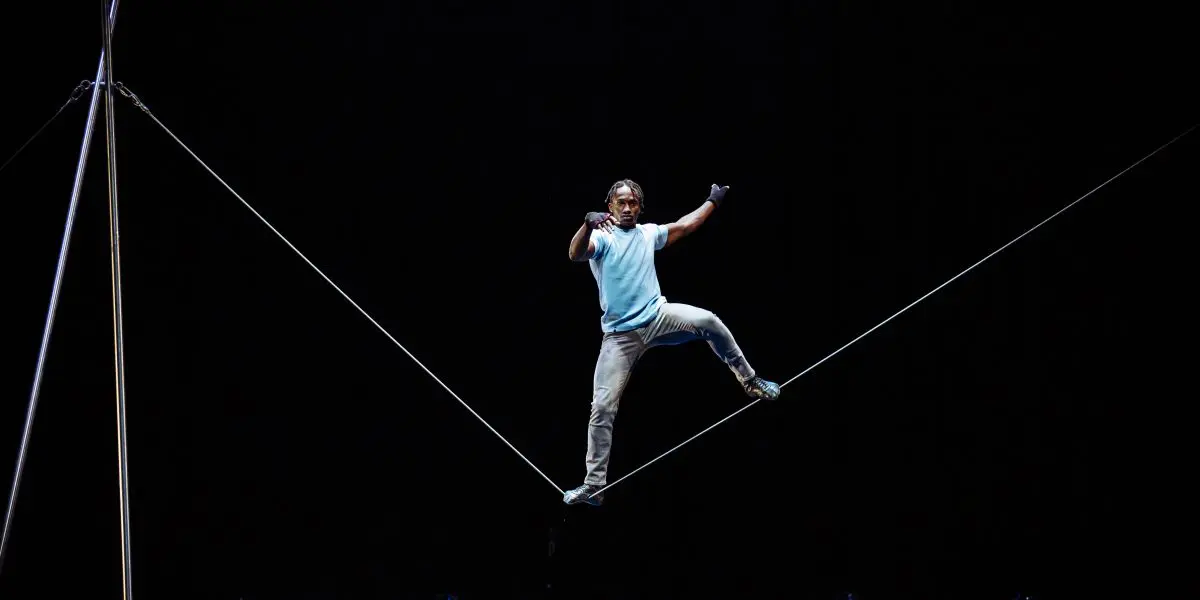 Antino Pansa, en chemise bleue, se tient en équilibre sur une corde raide en hauteur, sur un fond sombre, les bras levés pour l'équilibre et une jambe pliée.