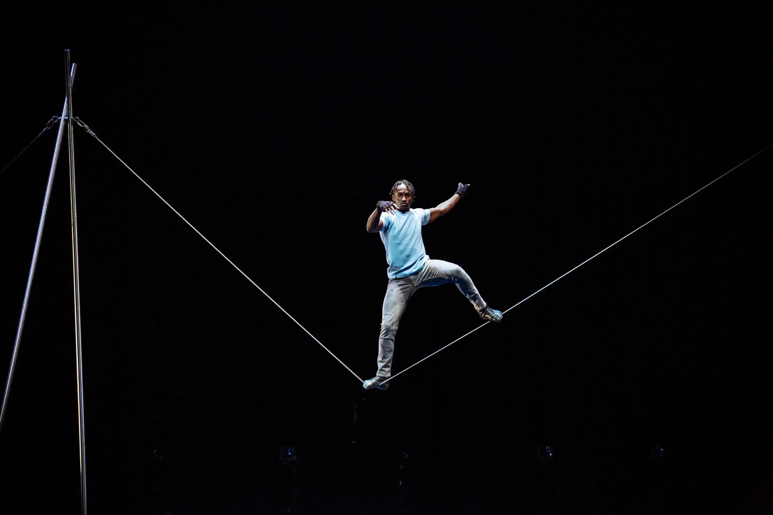 Antino Pansa, en chemise bleue, se tient en équilibre sur une corde raide en hauteur, sur un fond sombre, les bras levés pour l'équilibre et une jambe pliée.