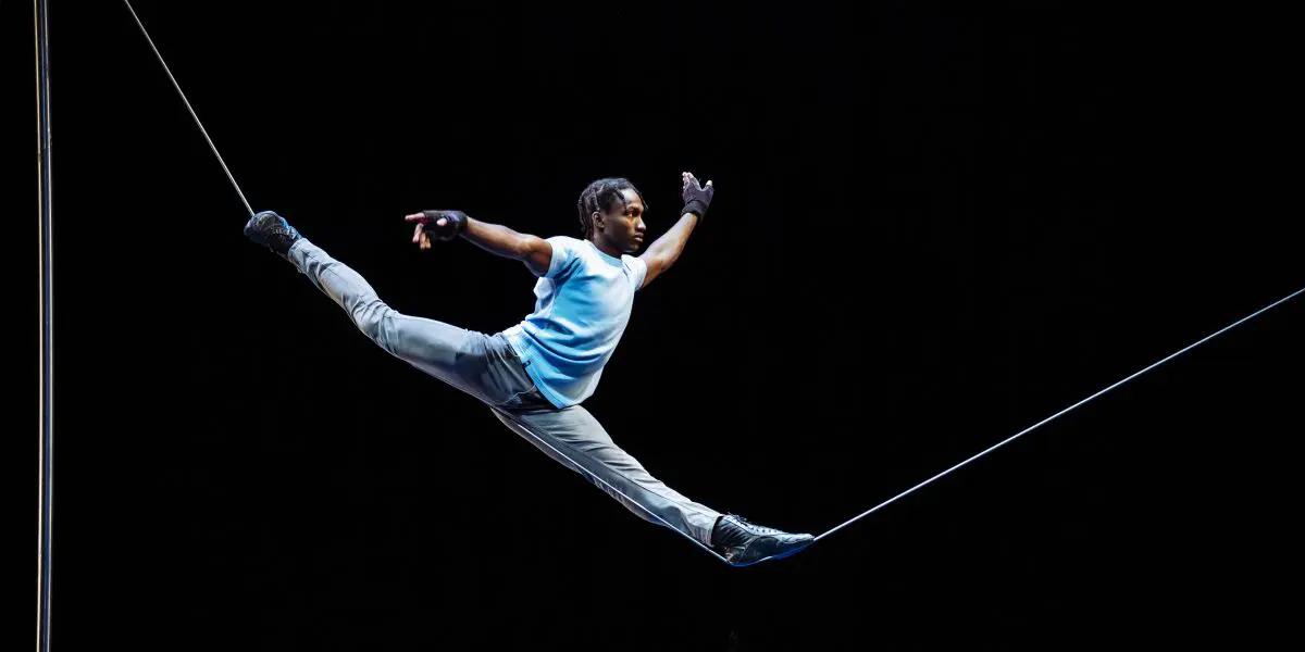 Antino Pansa, vêtu d'une chemise bleue et d'un pantalon gris, se tient en équilibre sur une corde raide, les bras tendus, sur un fond sombre, au cours d'un numéro de cirque ou d'acrobatie impressionnant.