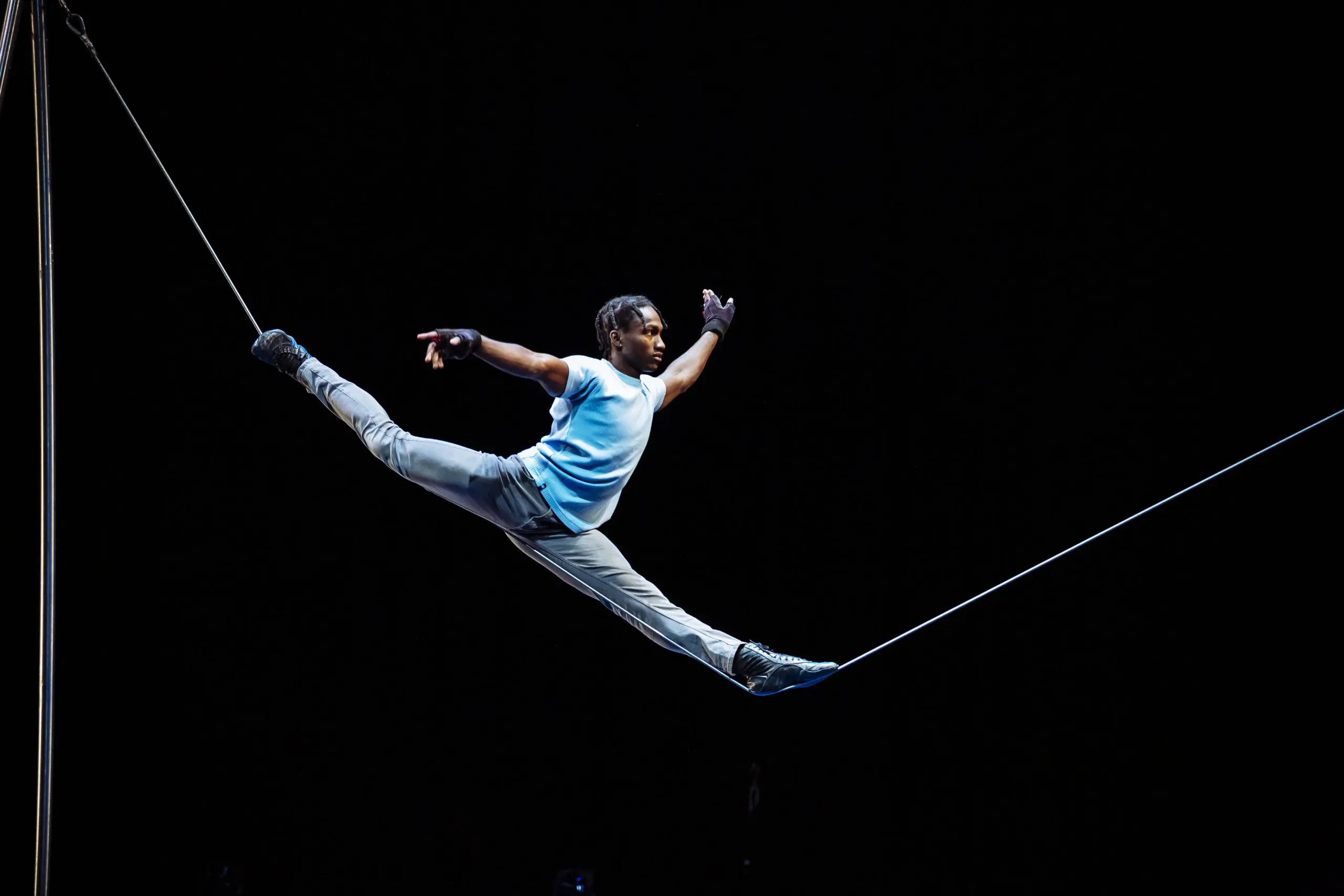 Antino Pansa, vêtu d'une chemise bleue et d'un pantalon gris, se tient en équilibre sur une corde raide, les bras tendus, sur un fond sombre, au cours d'un numéro de cirque ou d'acrobatie impressionnant.