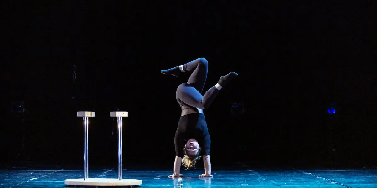 Amie Patching, vêtue de noir, effectue un équilibre sur les mains avec les jambes pliées sur une scène, avec deux plates-formes rondes sur de grands supports à proximité, sur un fond sombre.