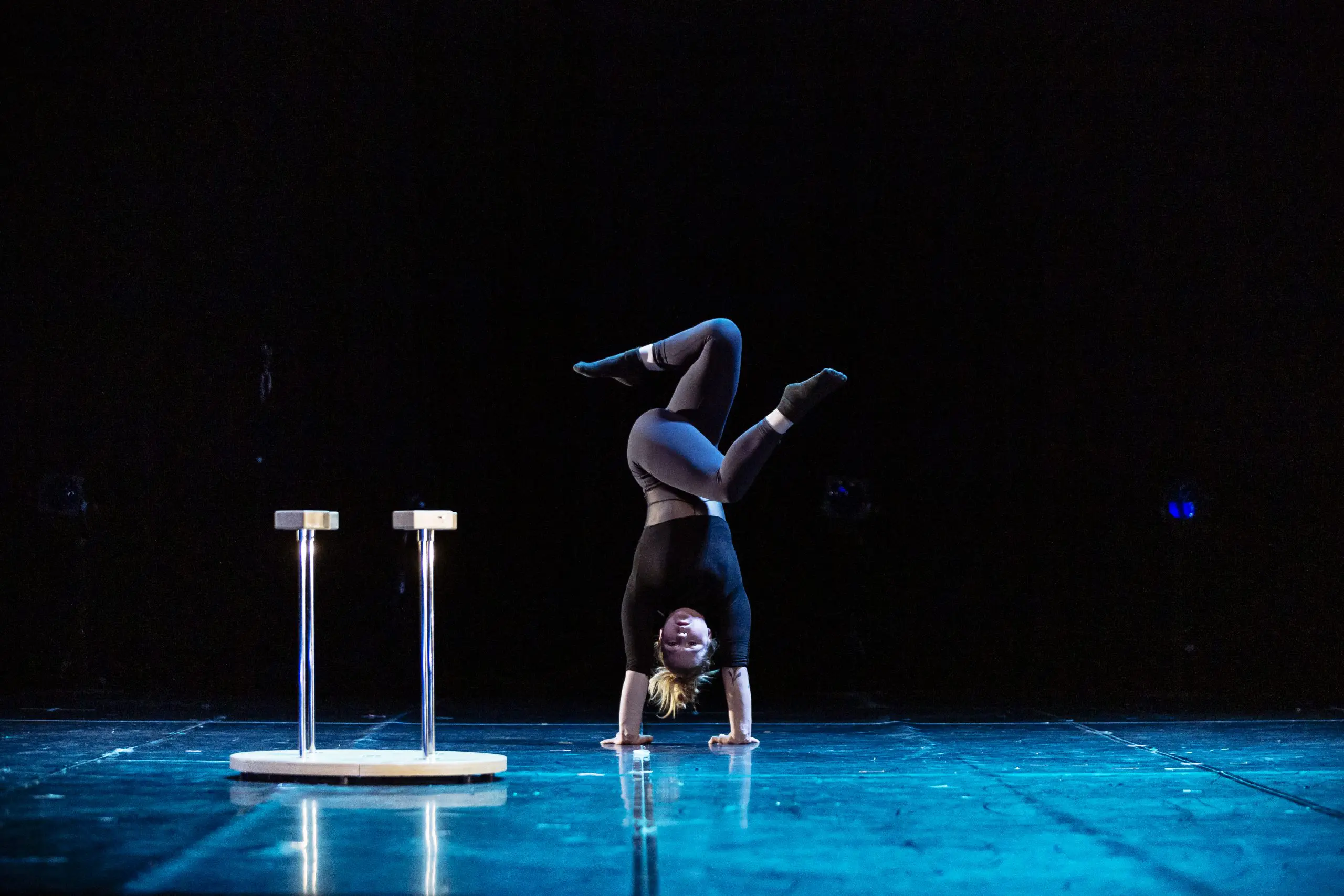 Amie Patching, vêtue de noir, effectue un équilibre sur les mains avec les jambes pliées sur une scène, avec deux plates-formes rondes sur de grands supports à proximité, sur un fond sombre.