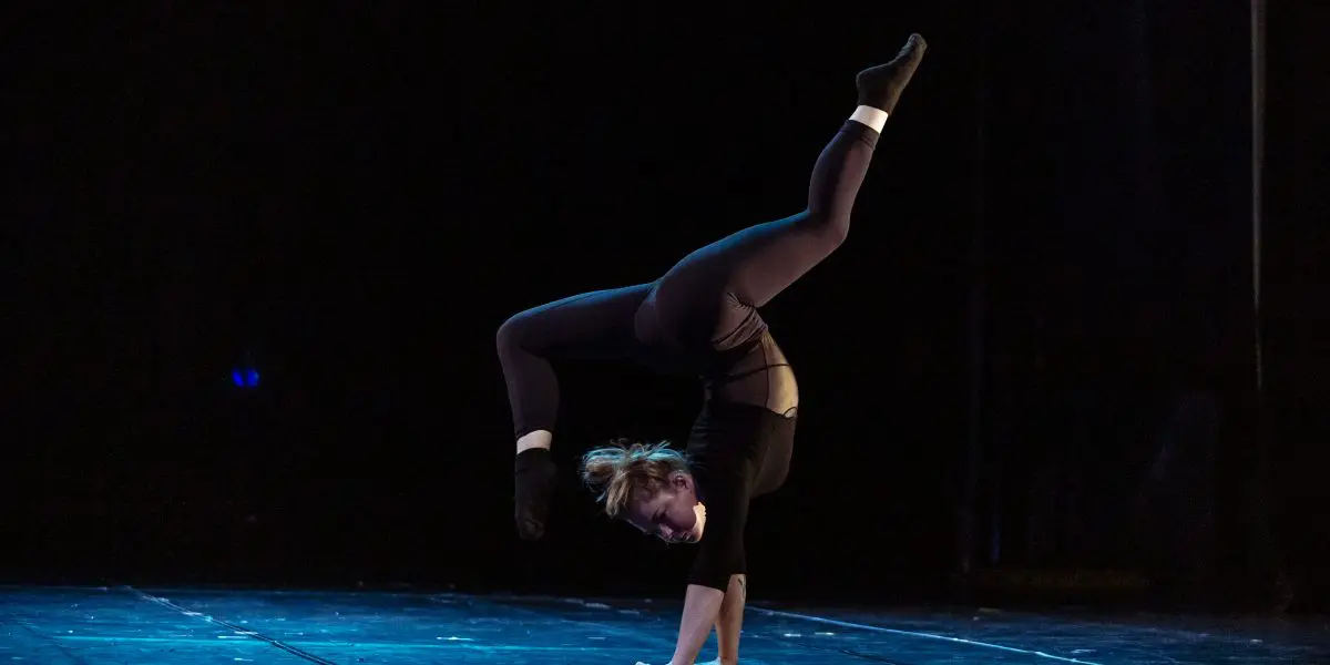 Amie Patching, vêtue de noir, exécute sur scène un remarquable poirier à une main, une jambe pliée et l'autre tendue vers le haut, éclairée par un éclairage de scène spectaculaire sur une toile de fond sombre.