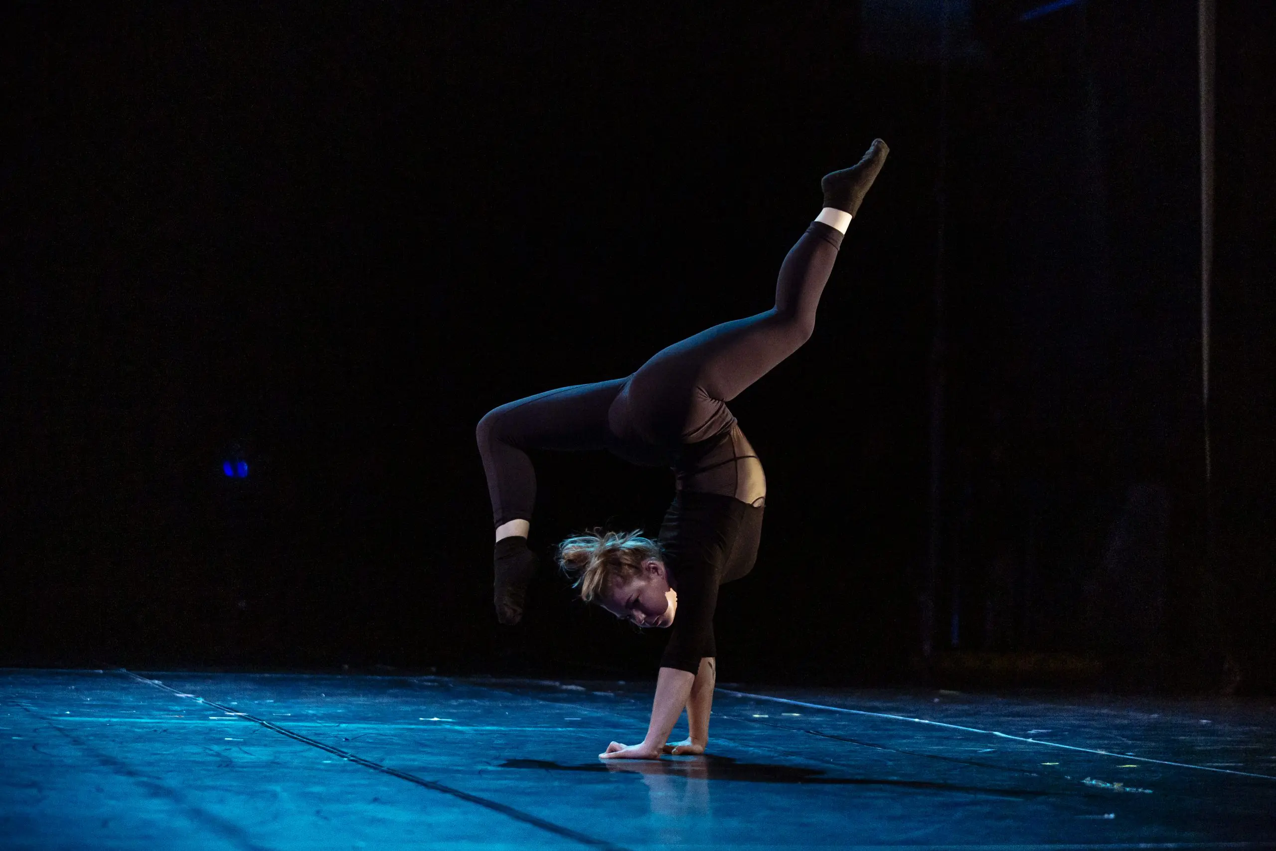Amie Patching, vêtue de noir, exécute sur scène un remarquable poirier à une main, une jambe pliée et l'autre tendue vers le haut, éclairée par un éclairage de scène spectaculaire sur une toile de fond sombre.