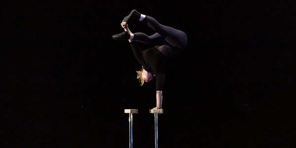 Amie Patching, une contorsionniste, se tient en équilibre sur les mains au sommet de deux plates-formes surélevées, les jambes pliées au-dessus de la tête et les pieds se touchant, sur un fond sombre.