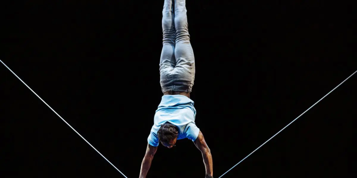 Antino Pansa réalise un équilibre sur une corde raide, la tête en bas, les deux mains agrippées à la corde. L'arrière-plan sombre met en évidence l'habileté et la concentration de l'acrobate.