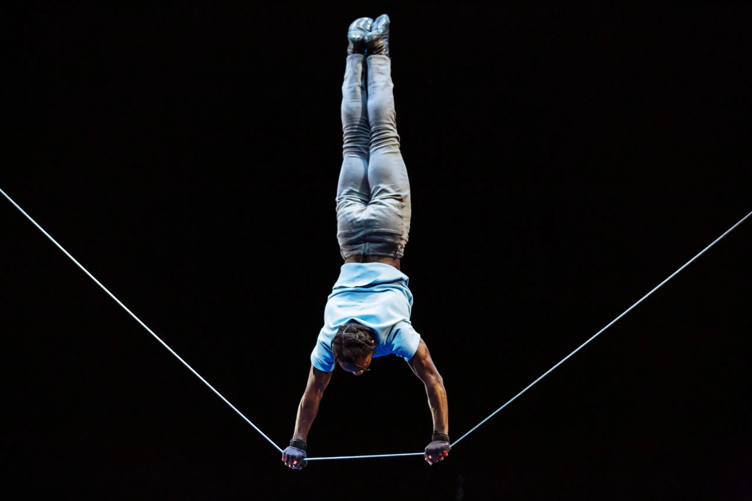 Antino Pansa réalise un équilibre sur une corde raide, la tête en bas, les deux mains agrippées à la corde. L'arrière-plan sombre met en évidence l'habileté et la concentration de l'acrobate.