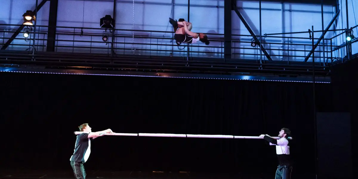 Deux artistes tiennent une longue perche à l'horizontale tandis que Naïka Aymon, une acrobate chevronnée, s'élance au-dessus d'eux en plein vol, dans une salle de spectacle moderne et lumineuse.