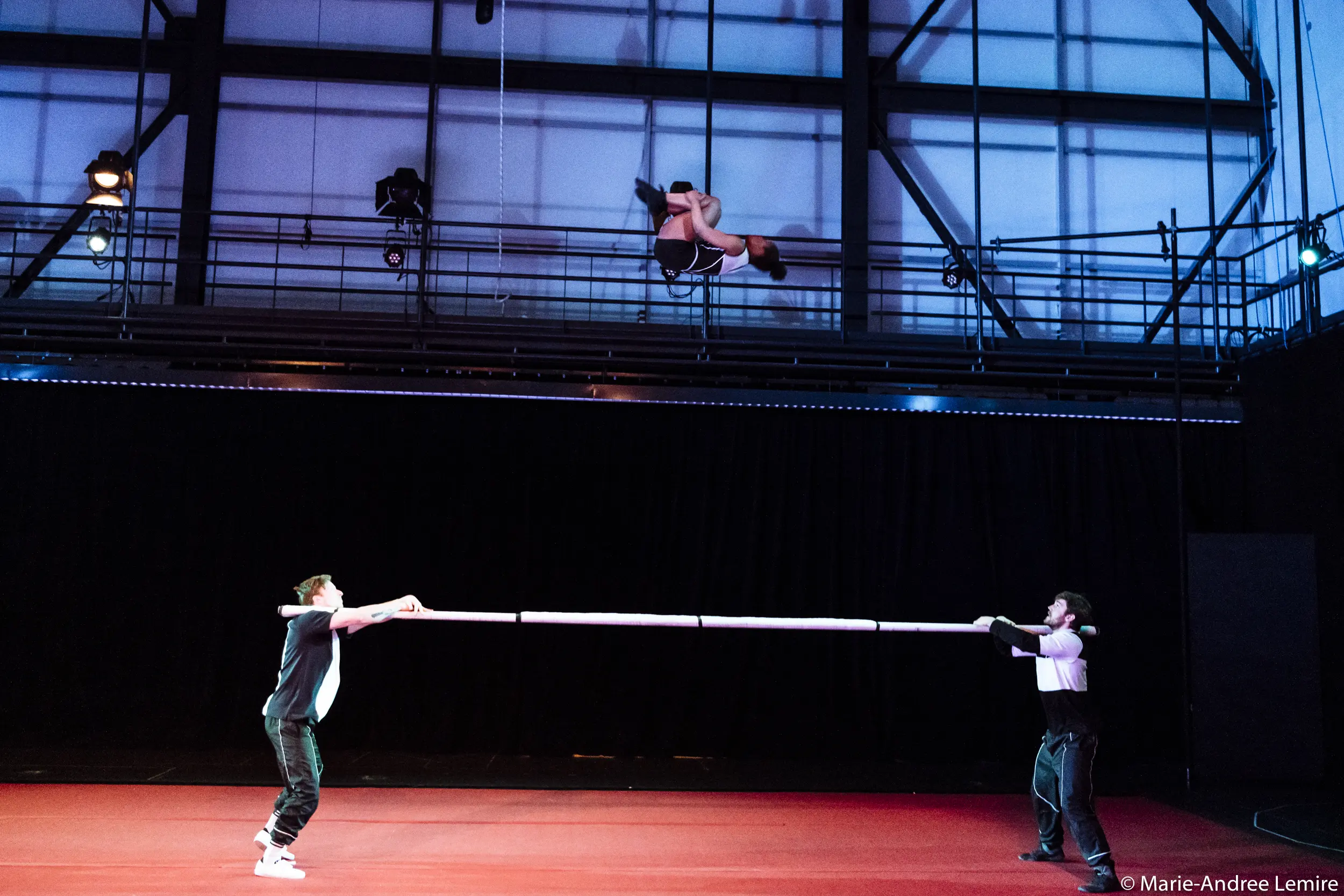 Deux artistes tiennent une longue perche à l'horizontale tandis que Naïka Aymon, une acrobate chevronnée, s'élance au-dessus d'eux en plein vol, dans une salle de spectacle moderne et lumineuse.