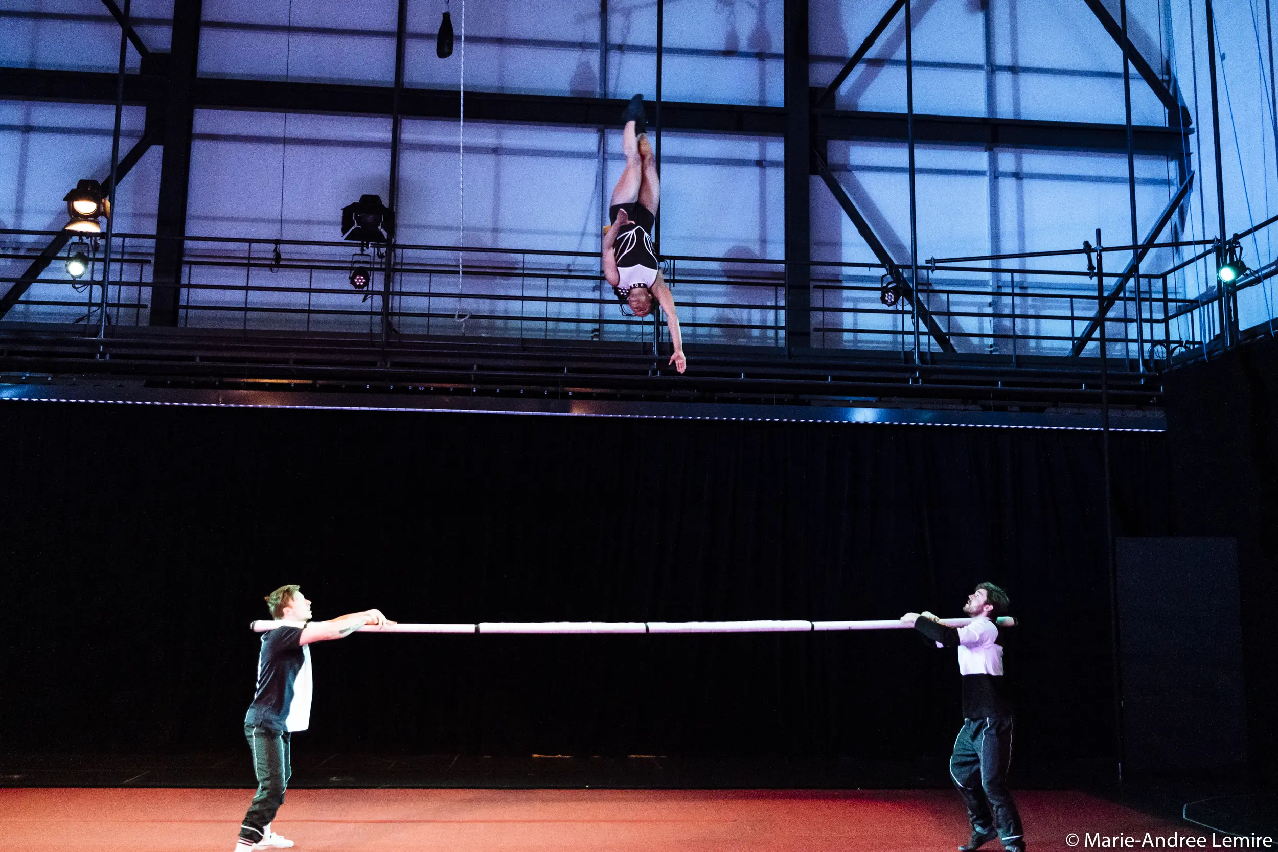 Un artiste est suspendu à l'envers à une corde en haut d'une scène, tandis que Naïka Aymon et une autre personne tiennent un tapis de sécurité sous eux dans un gymnase ou une salle de spectacle très éclairée.