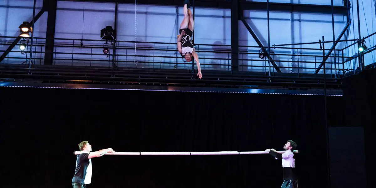 Un artiste est suspendu au plafond au-dessus d'une scène, la tête en bas, tandis que deux personnes au sol tiennent une longue perche horizontale sous elles, se préparant à un numéro d'acrobatie de Benjamin Wolgers.