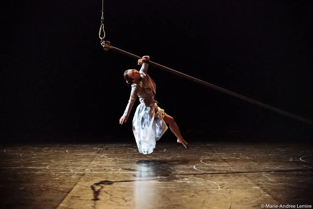 L'artiste Minji Kim, vêtue d'une robe fluide, se suspend gracieusement à une corde épaisse au-dessus d'une scène faiblement éclairée, un bras tendu et les jambes en équilibre dans les airs.