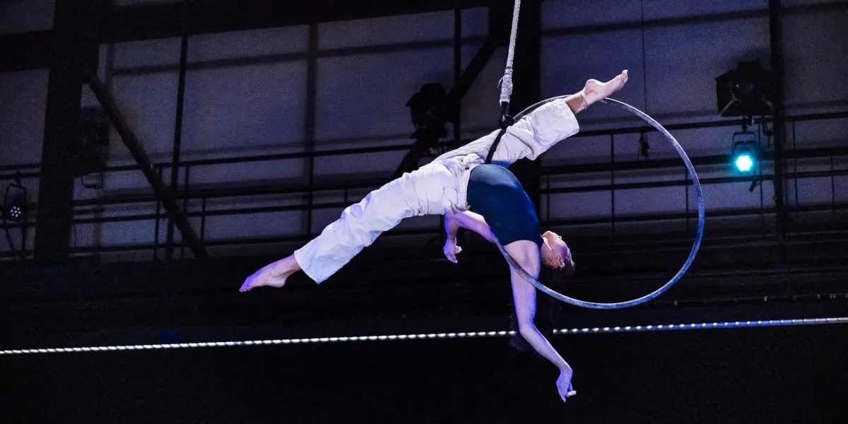 L'artiste aérienne Michelle Sargent exécute une pose acrobatique sur un cerceau suspendu, une jambe tendue vers le haut et l'autre vers l'extérieur, dans un décor intérieur dramatique mis en valeur par un éclairage de scène époustouflant.