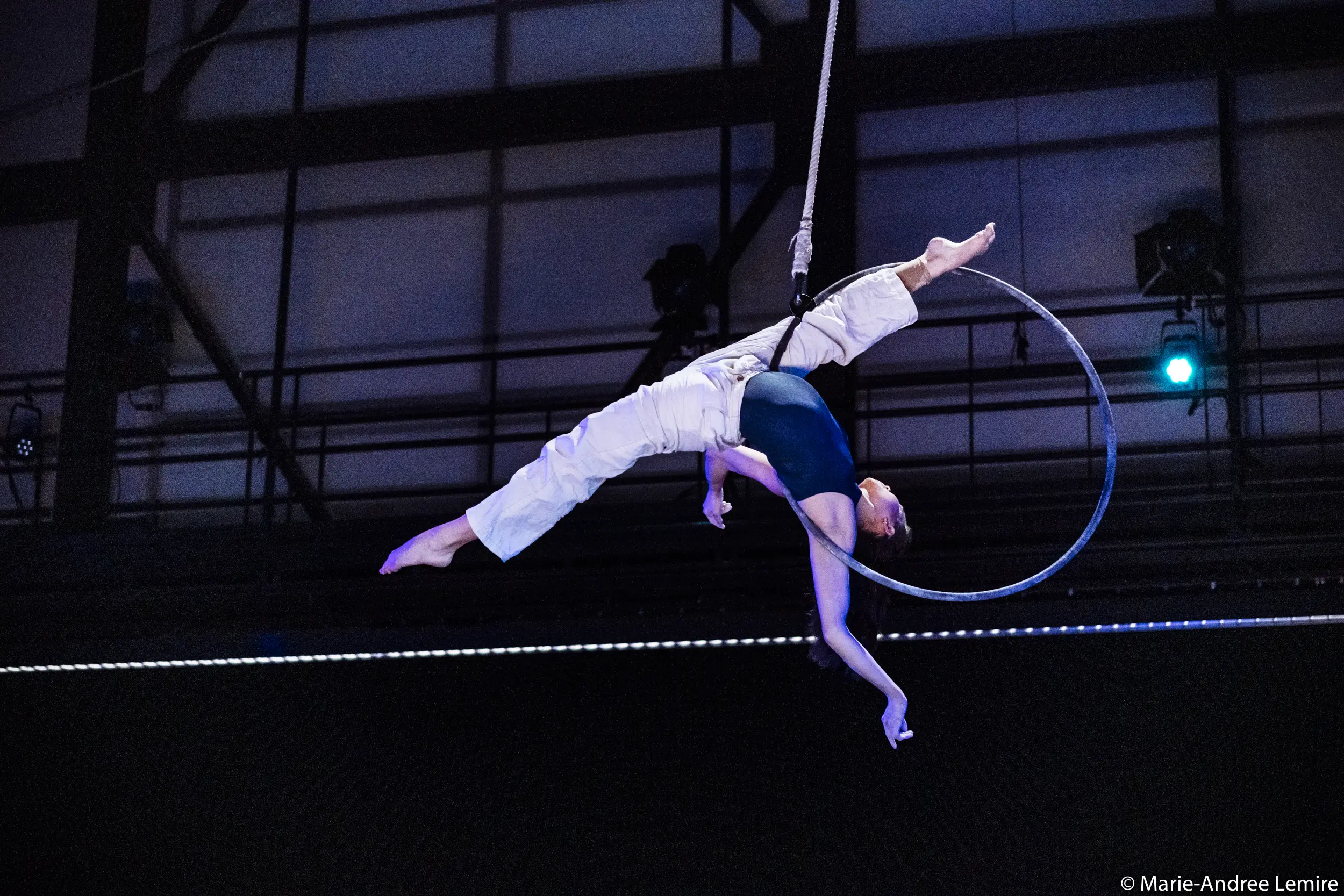 L'artiste aérienne Michelle Sargent exécute une pose acrobatique sur un cerceau suspendu, une jambe tendue vers le haut et l'autre vers l'extérieur, dans un décor intérieur dramatique mis en valeur par un éclairage de scène époustouflant.
