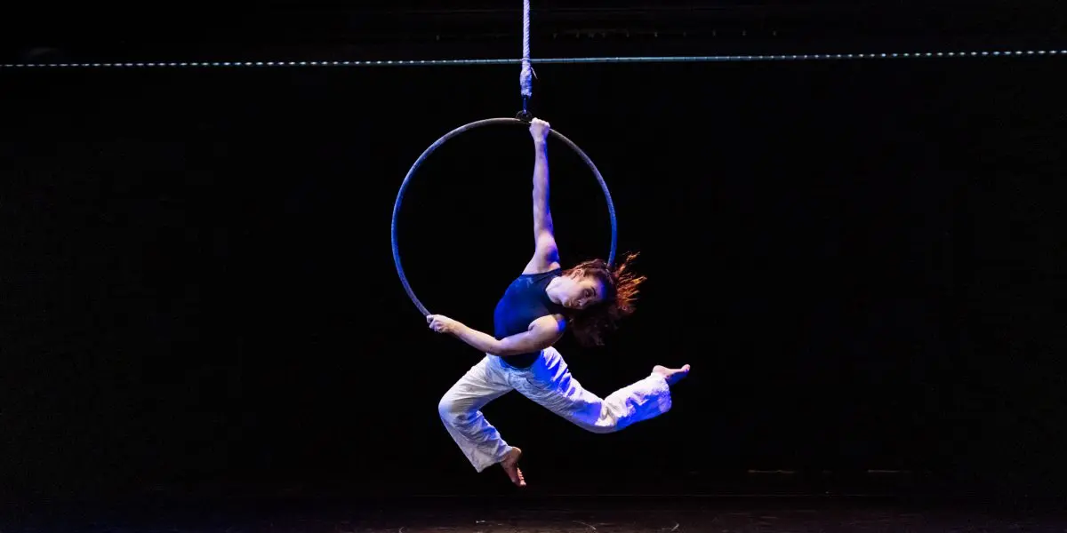 Dans un théâtre sombre, l'artiste aérienne Michelle Sargent se suspend gracieusement à un cerceau suspendu, se tenant à un bras tout en arquant son corps et en étendant ses jambes dans les airs. Elle porte un haut bleu marine et un pantalon blanc.