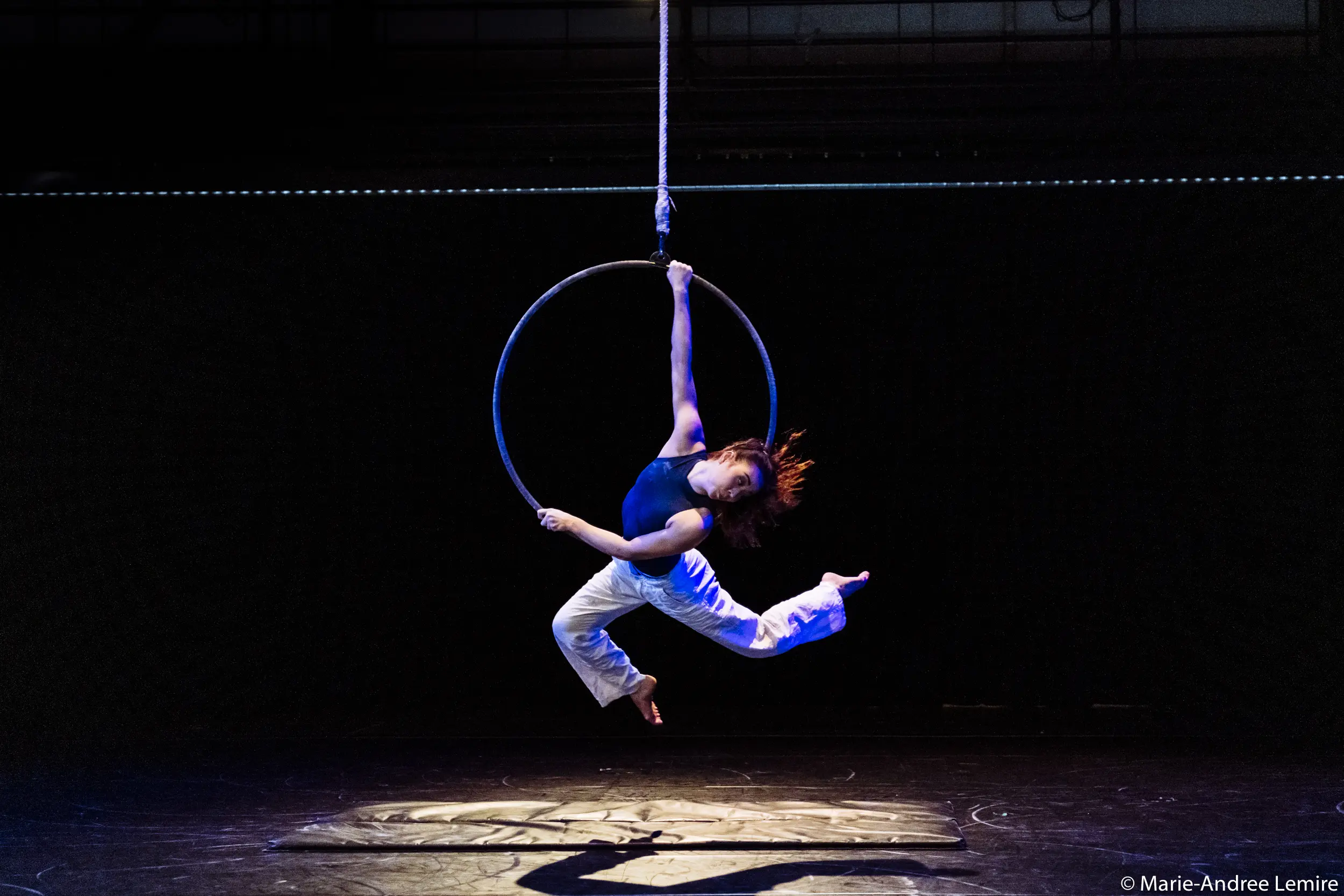 Dans un théâtre sombre, l'artiste aérienne Michelle Sargent se suspend gracieusement à un cerceau suspendu, se tenant à un bras tout en arquant son corps et en étendant ses jambes dans les airs. Elle porte un haut bleu marine et un pantalon blanc.