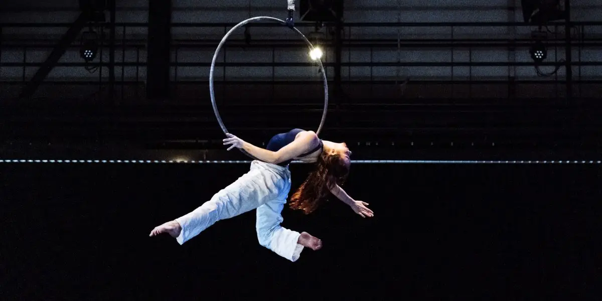 L'artiste aérienne Michelle Sargent, aux longs cheveux roux et au pantalon blanc, se suspend gracieusement à un cerceau, arquant son dos et étendant ses bras et ses jambes sur un fond sombre.
