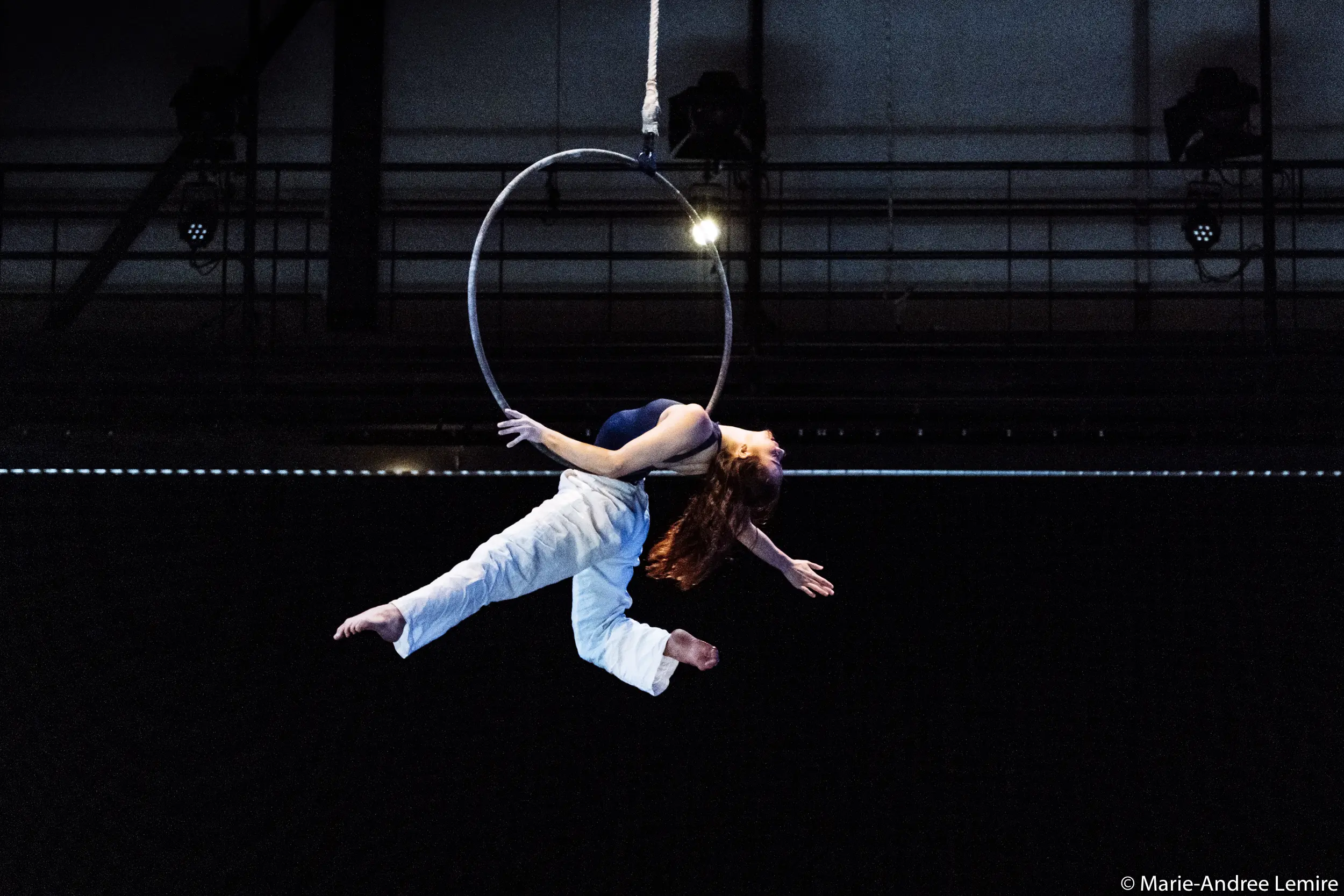 L'artiste aérienne Michelle Sargent, aux longs cheveux roux et au pantalon blanc, se suspend gracieusement à un cerceau, arquant son dos et étendant ses bras et ses jambes sur un fond sombre.