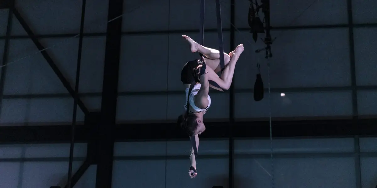 L'artiste aérienne Clara Laurent prend la pose à l'envers sur des soies, suspendue en hauteur contre une grande fenêtre à cadre métallique faiblement éclairée.