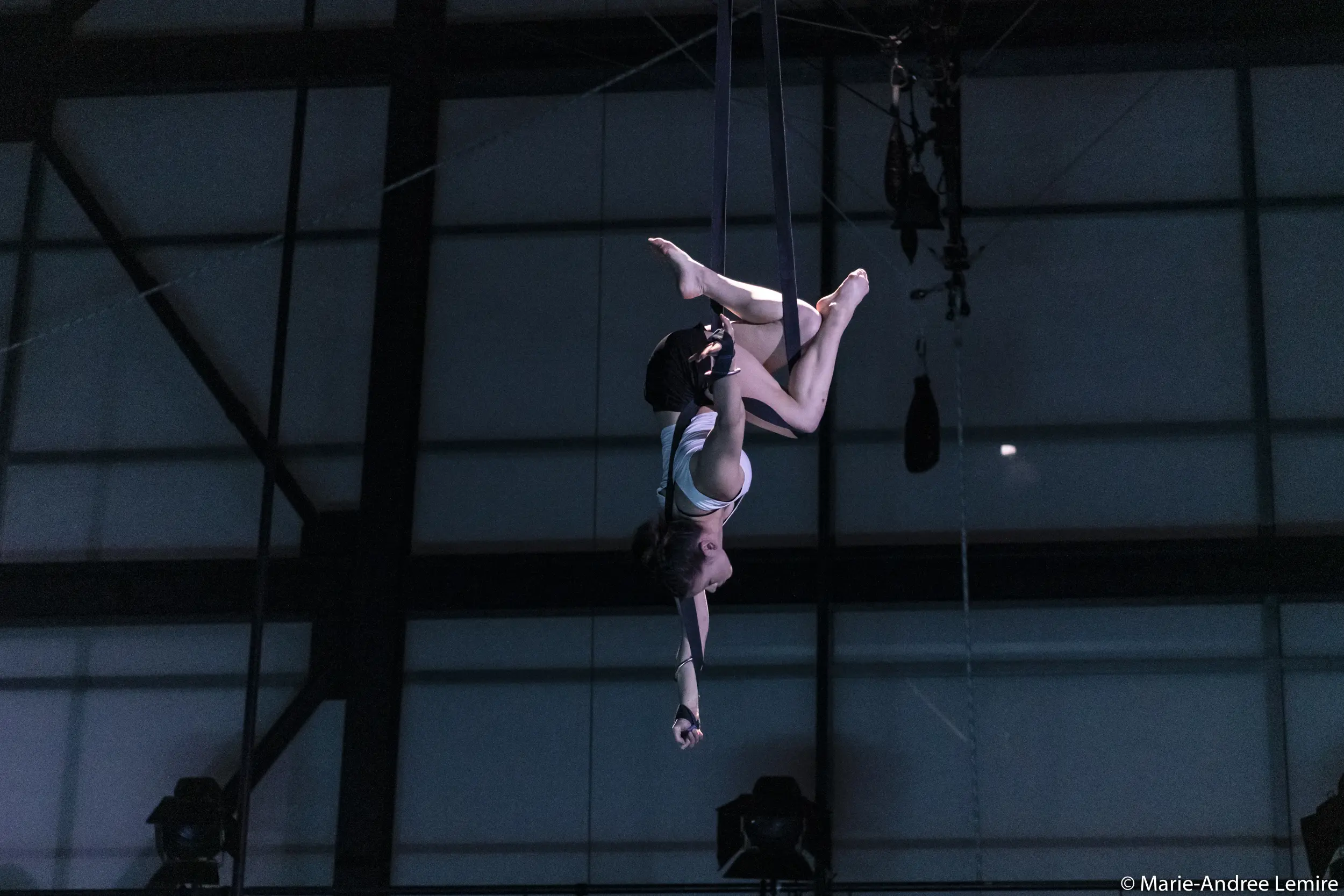 L'artiste aérienne Clara Laurent prend la pose à l'envers sur des soies, suspendue en hauteur contre une grande fenêtre à cadre métallique faiblement éclairée.