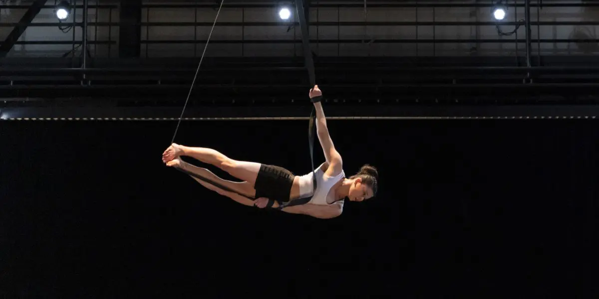 Clara Laurent, une artiste aérienne en tenue de sport, exécute une pose horizontale en étant suspendue par des sangles, surélevée au-dessus d'une scène sombre éclairée par des lumières.