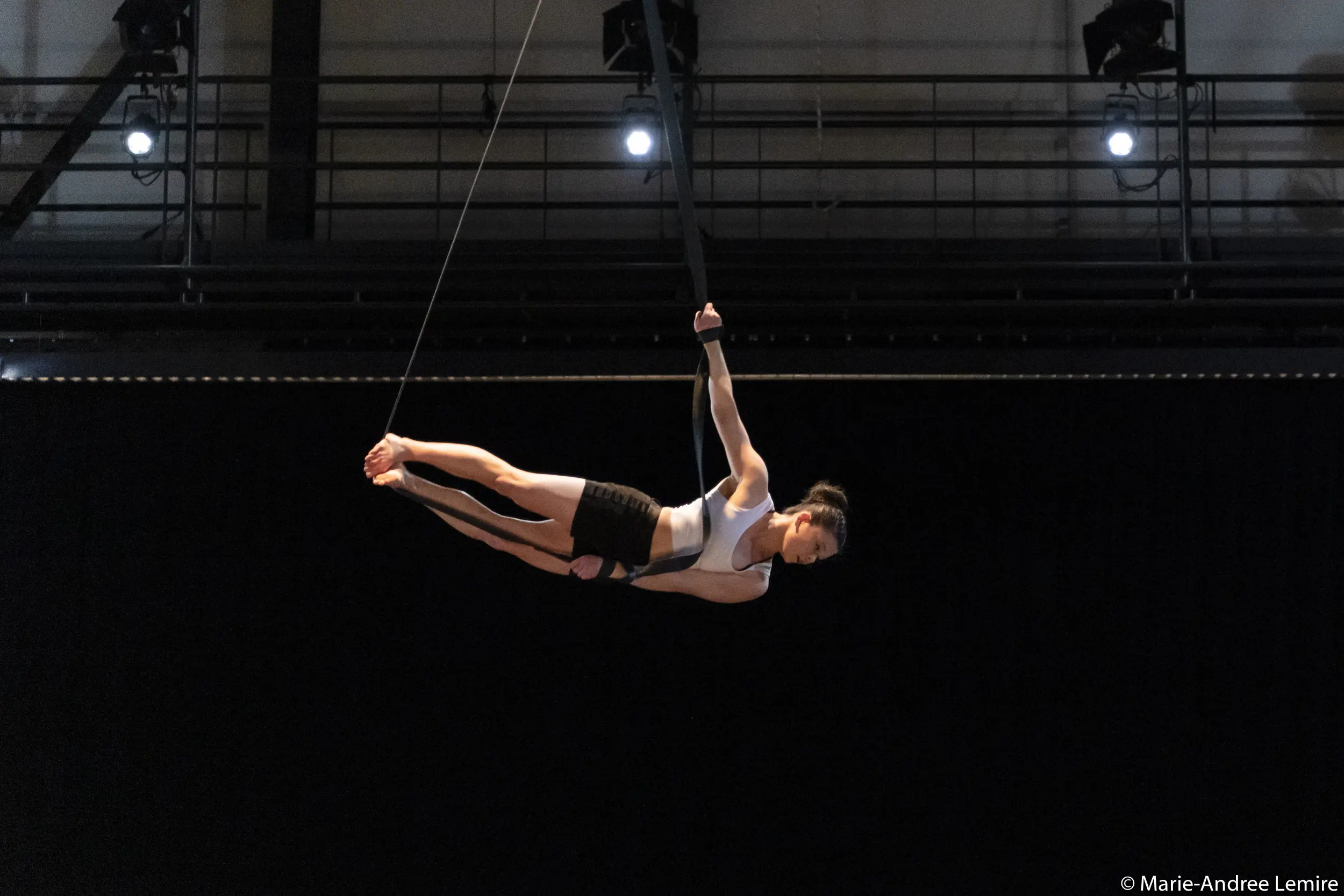 Clara Laurent, une artiste aérienne en tenue de sport, exécute une pose horizontale en étant suspendue par des sangles, surélevée au-dessus d'une scène sombre éclairée par des lumières.