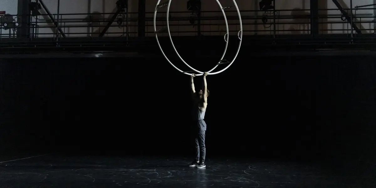 Elijah Kirk se tient sur une scène sombre, les bras levés pour tenir deux grands anneaux métalliques à la verticale au-dessus de sa tête, éclairés par un projecteur.