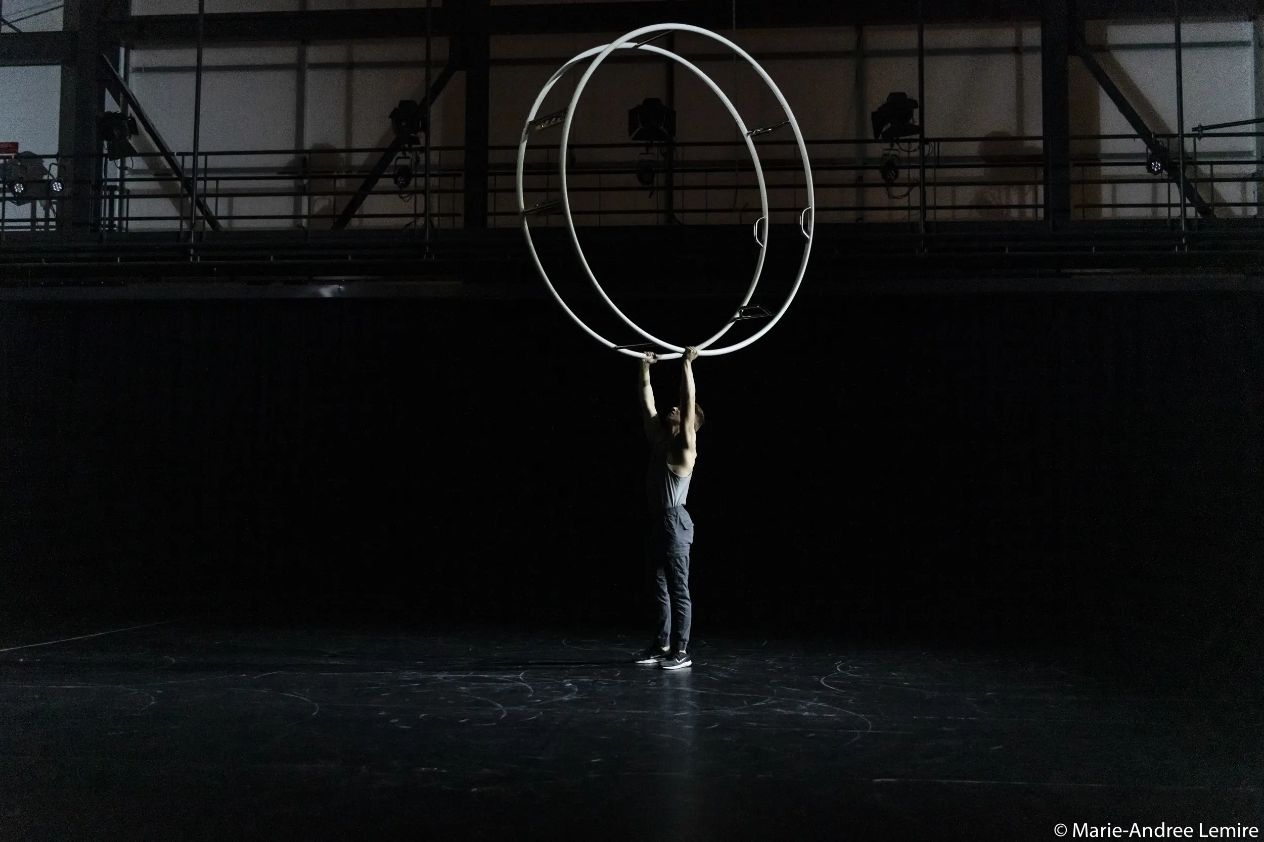 Elijah Kirk se tient sur une scène sombre, les bras levés pour tenir deux grands anneaux métalliques à la verticale au-dessus de sa tête, éclairés par un projecteur.