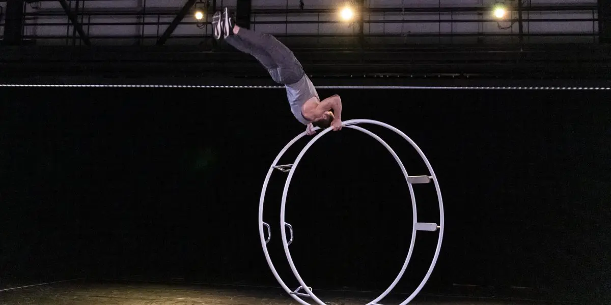 Un artiste, Elijah Kirk, se tient en équilibre à l'envers sur une grande roue métallique sous les lumières de la scène, tenant la roue à deux mains tandis que son corps s'étend verticalement dans une pose acrobatique dramatique.