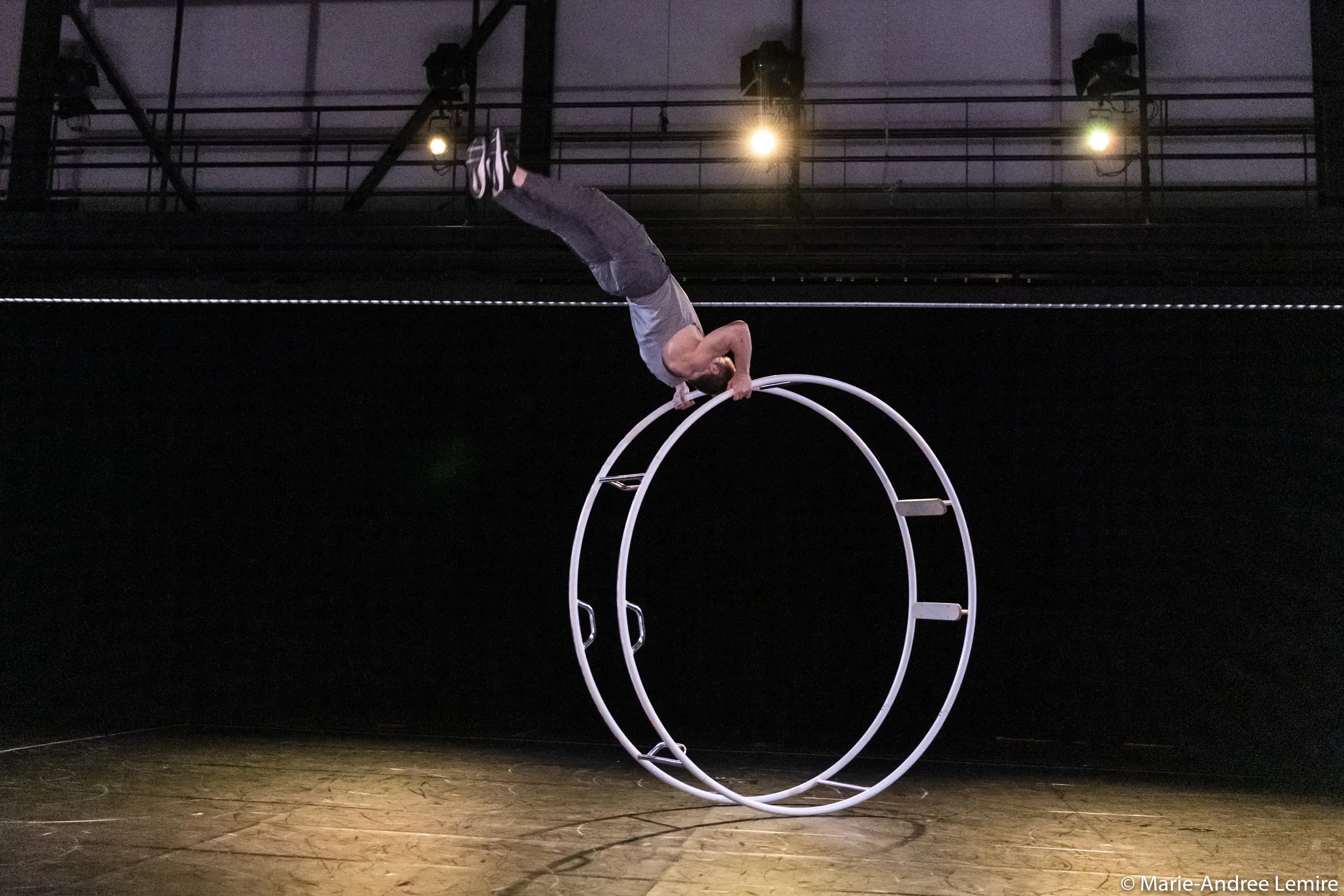 Un artiste, Elijah Kirk, se tient en équilibre à l'envers sur une grande roue métallique sous les lumières de la scène, tenant la roue à deux mains tandis que son corps s'étend verticalement dans une pose acrobatique dramatique.