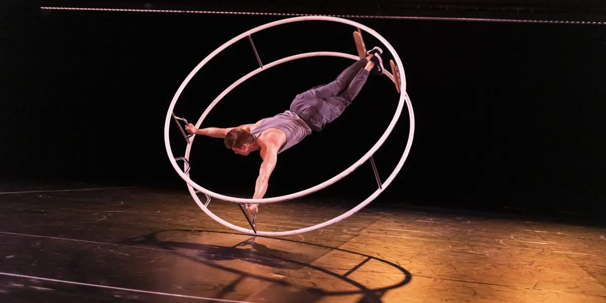 Elijah Kirk se tient en équilibre sur une grande roue Cyr blanche, saisissant le cadre avec les mains et les pieds, tout en étant suspendu horizontalement au-dessus d'une scène sous un éclairage chaleureux.