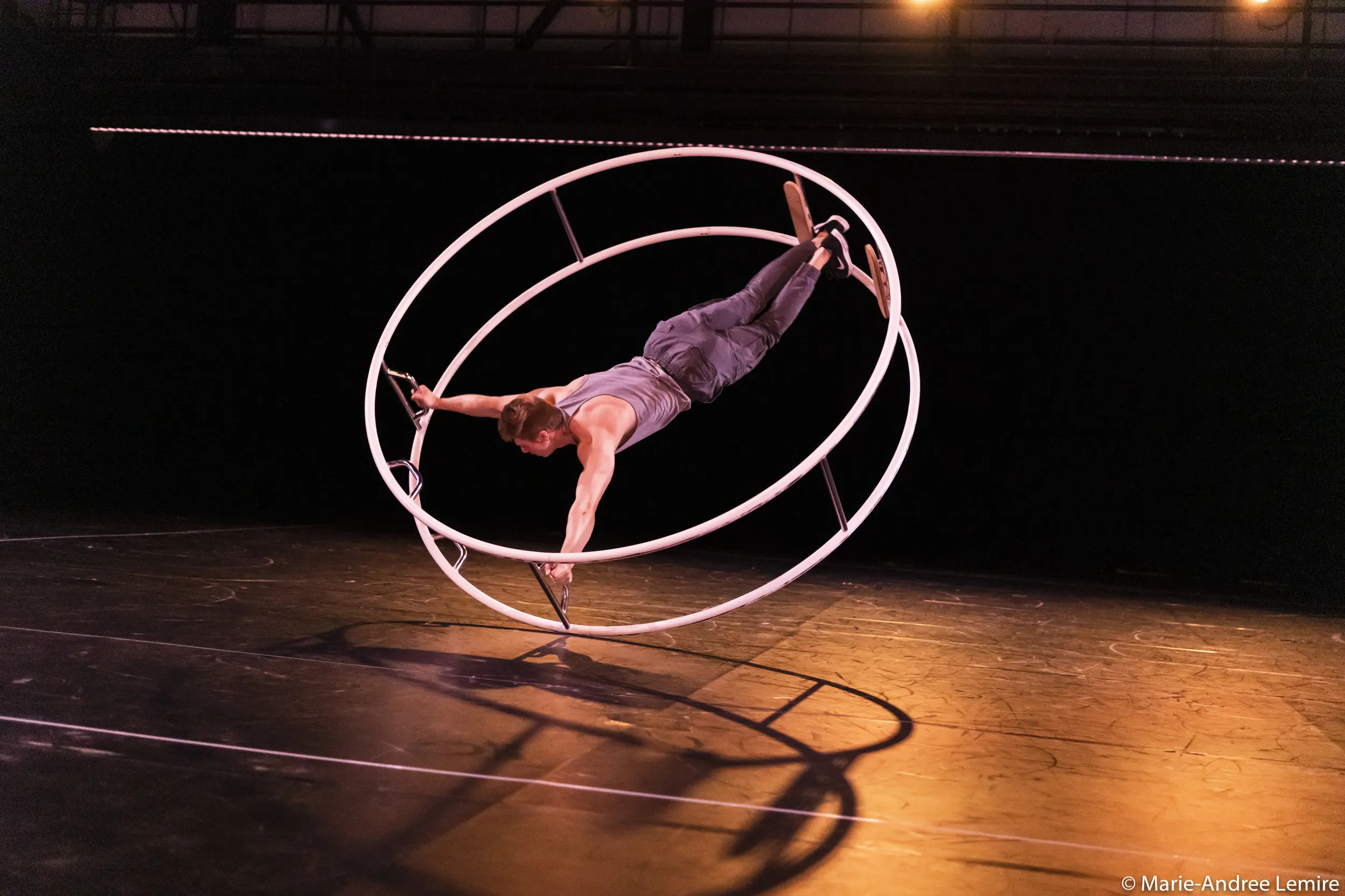 Elijah Kirk se tient en équilibre sur une grande roue Cyr blanche, saisissant le cadre avec les mains et les pieds, tout en étant suspendu horizontalement au-dessus d'une scène sous un éclairage chaleureux.