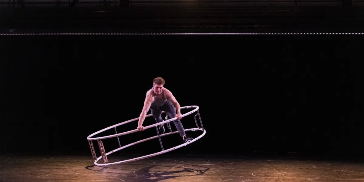 Elijah Kirk se tient en équilibre sur une grande roue de cyr en métal, l'inclinant de biais sur une scène faiblement éclairée par des projecteurs, sur un fond sombre.