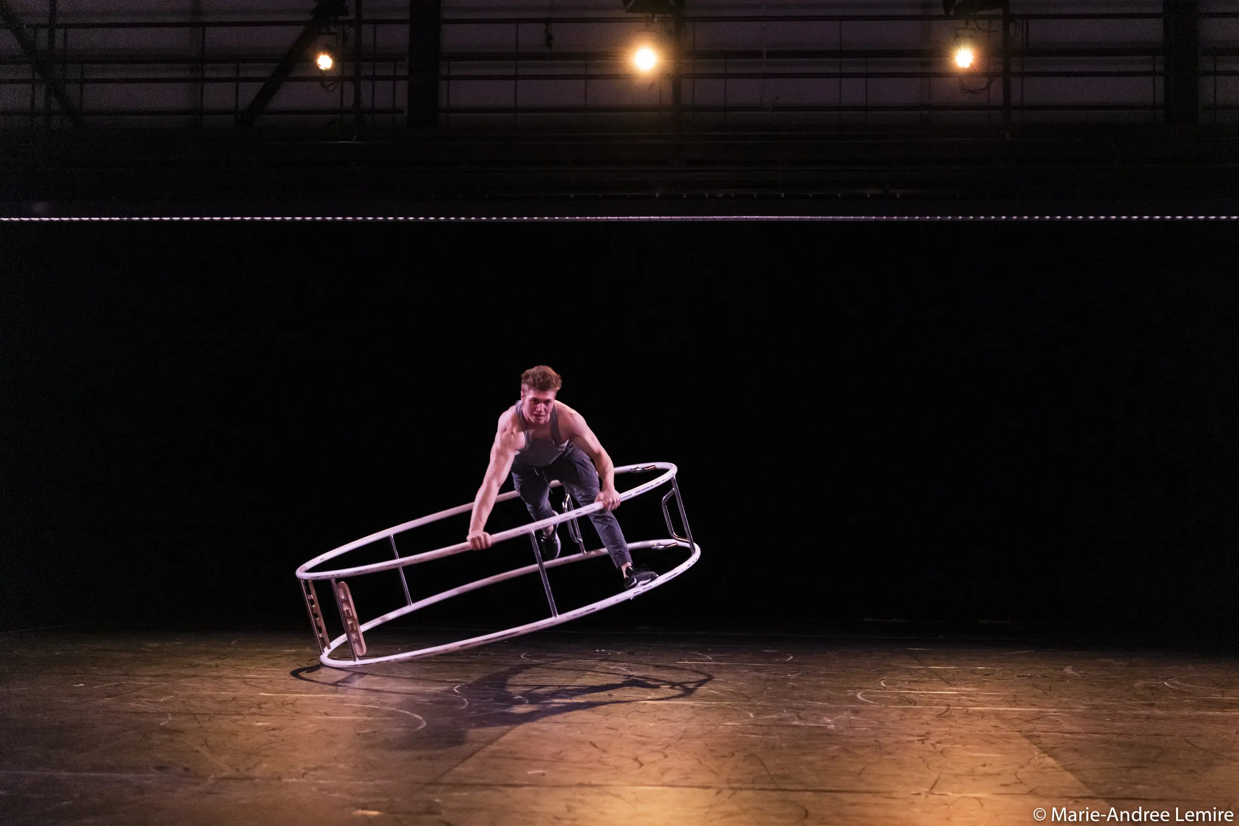 Elijah Kirk se tient en équilibre sur une grande roue de cyr en métal, l'inclinant de biais sur une scène faiblement éclairée par des projecteurs, sur un fond sombre.