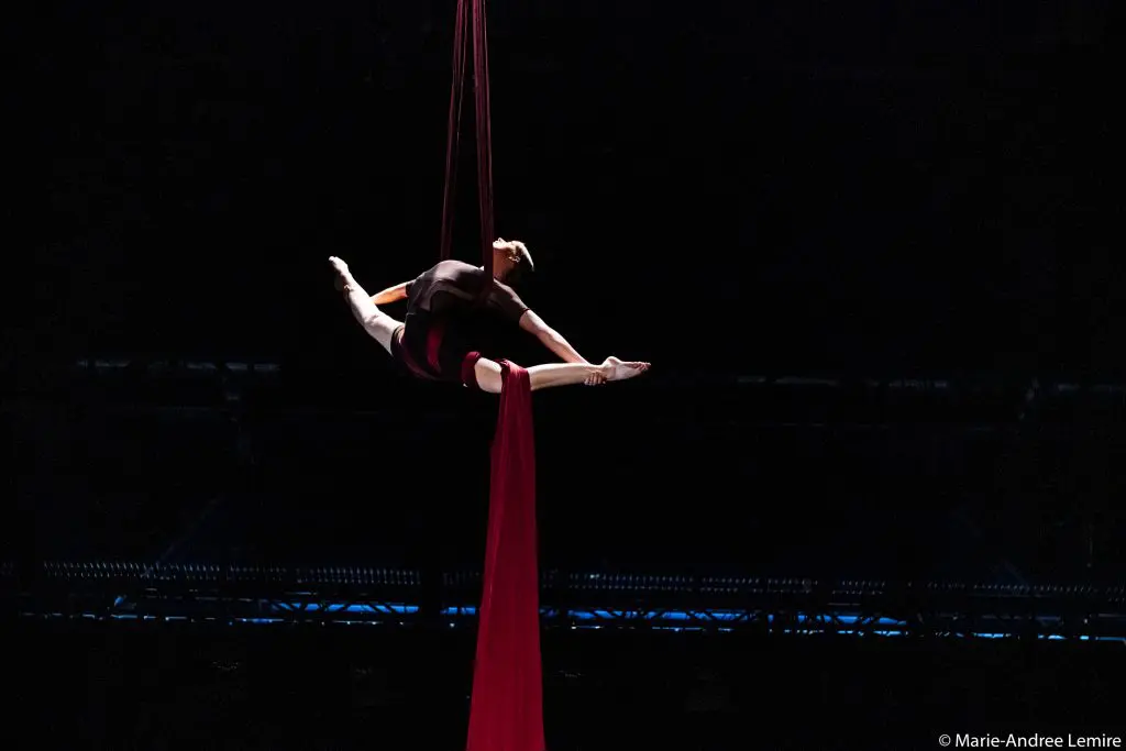 L'artiste aérienne Jasmine Gagné-Morin est suspendue dans les airs par des soies rouges, les bras et les jambes étendus dans une pose fendue sur un fond sombre. Les lumières de la scène éclairent faiblement la scène.