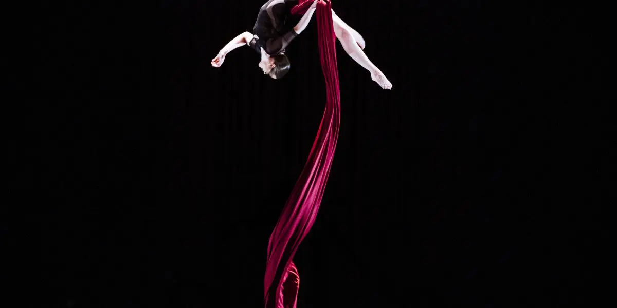Jasmine Gagné-Morin, une artiste aérienne en soie, vêtue d'une tenue noire, est suspendue la tête en bas, posant gracieusement dans les airs avec un tissu de soie rouge qui spirale sous elle, sur un fond noir.