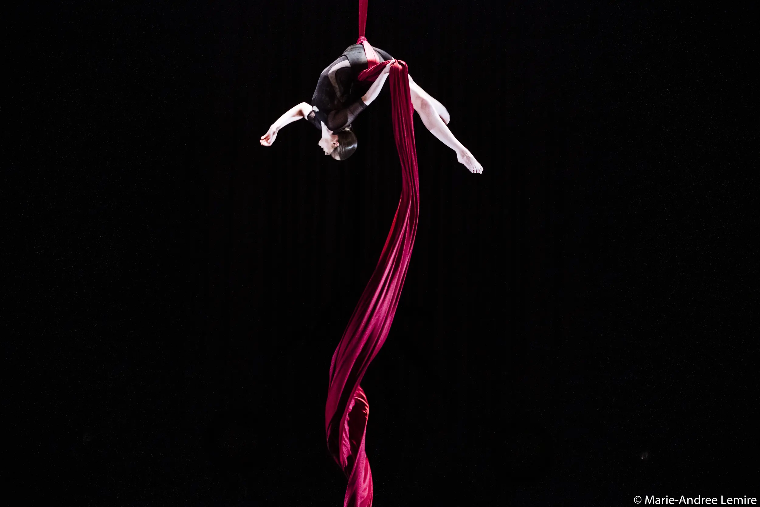 Jasmine Gagné-Morin, une artiste aérienne en soie, vêtue d'une tenue noire, est suspendue la tête en bas, posant gracieusement dans les airs avec un tissu de soie rouge qui spirale sous elle, sur un fond noir.