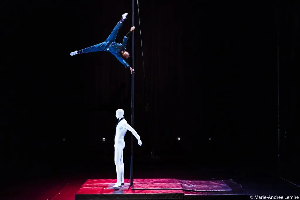 Une performeuse en bleu, peut-être Naïka Aymon, se tient en équilibre horizontal sur un poteau vertical au-dessus d'un tapis rouge, tandis qu'un mannequin blanc se tient en dessous d'eux, le tout sur un fond sombre.