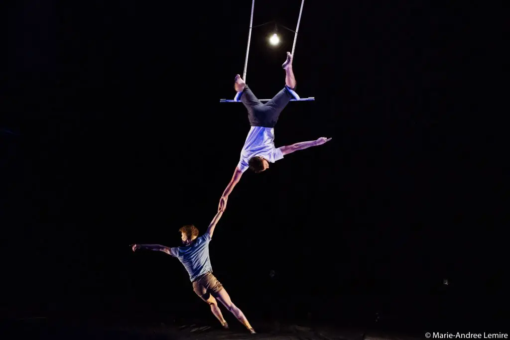 Deux trapézistes, Maxime Charron et Corentin Lemaître Auger, évoluent dans les airs ; l'un est suspendu à une barre de trapèze, la tête en bas, et tend la main vers l'autre acrobate, tous deux suspendus sur un fond sombre.