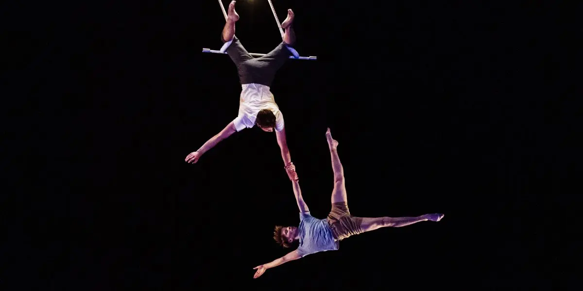 Deux acrobates, Maxime Charron et Corentin Lemaître Auger, sont suspendus en l'air sur un trapèze, l'un à l'envers et tenant la main de l'autre, faisant preuve d'un équilibre et d'une force remarquables sur un fond sombre.